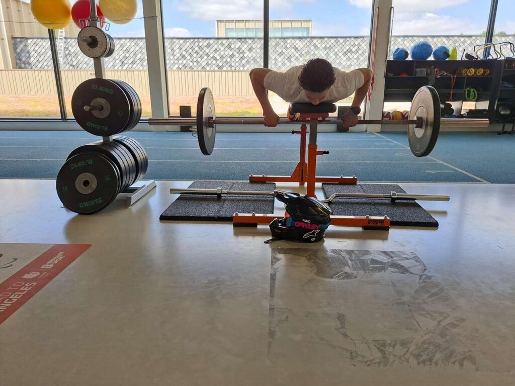 Motorcrosser Dean Gregoire doet een rugoefening over een trainingsbank in een sportschool in Eindhoven, met zijn crosshelm op de voorgrond.