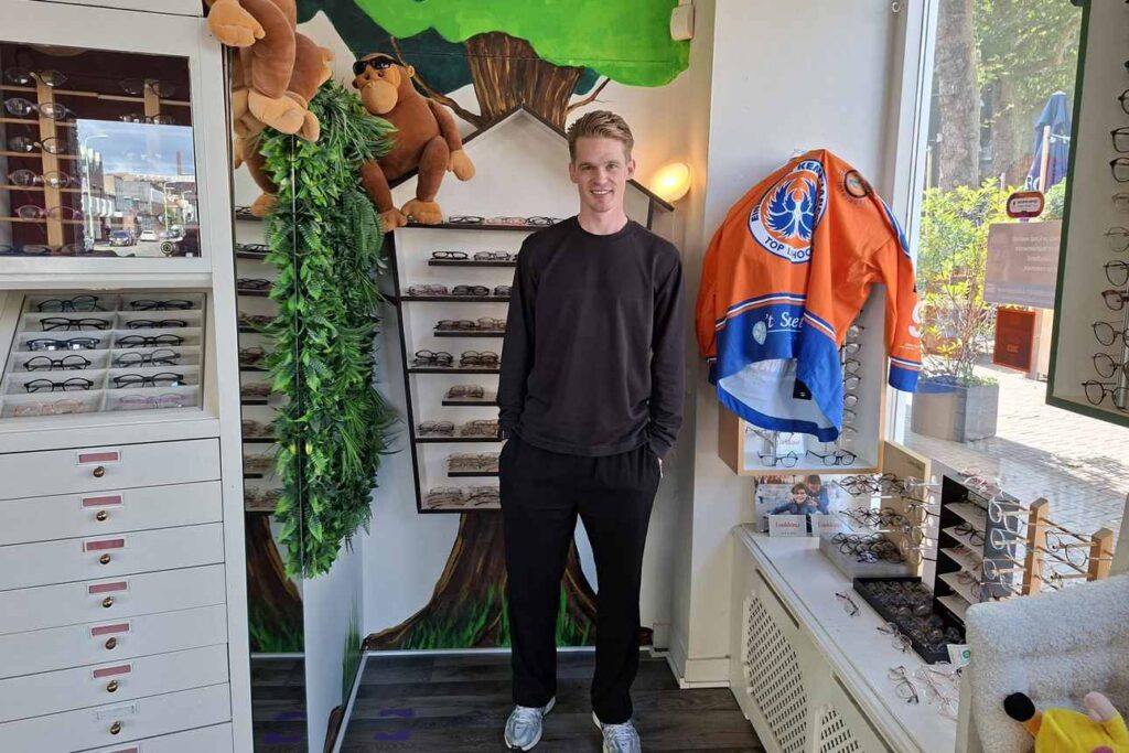 Mike Verschuren, captain van de Kemphanen, staat in een brillenwinkel voor een muurschildering van een boom. Rechts van hem hangt een oranje-blauw shirt van de Kemphanen.