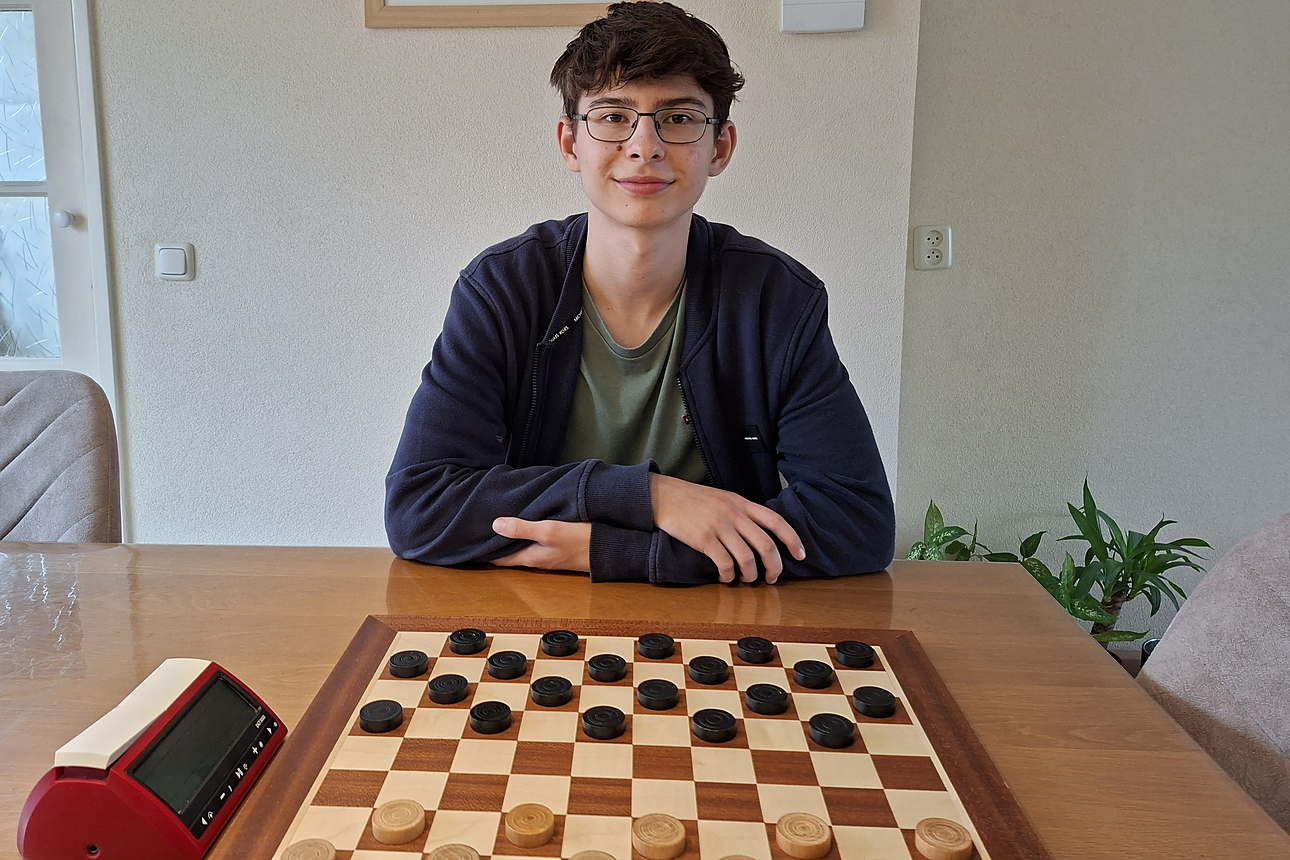 Alex Ovsyanko zit thuis aan de eettafel achter een opgezet dambord, met een damklok links naast het bord.