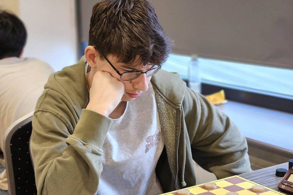 Alex Ovsyanko denkt na boven het dambord tijdens een damtoernooi, zijn hoofd rustend op zijn vuist.